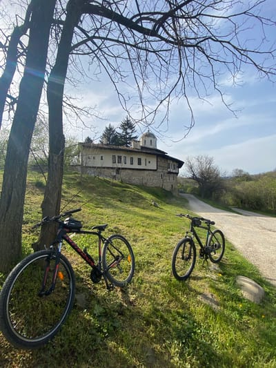 The image shows a landscape in Pchelishte, Bulgaria. A stone building with a dome is visible in the background, possibly a monastery or church. Two mountain bikes are lying on the grass, one black and one black and red. A paved road leads past the building. A bare tree is in the foreground on the left. The sky is blue with wispy clouds. The scene is set during daylight.