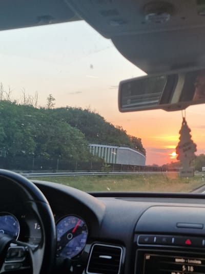 The image is taken from the interior of a moving car, looking forward through the windshield. The dashboard is visible in the foreground, with the steering wheel on the left and a speedometer displaying a speed of approximately 100 km/h on the right. Outside the car, a sunset is occurring with the sun low on the horizon, casting an orange glow across the sky. The landscape includes a tree-covered hill to the left and a white barrier structure. In the distance, a car is visible on the road. A tree-shaped air freshener hangs from the rearview mirror. The scene is likely the M7 motorway in Balatonszemes, Hungary, known for its scenic sunsets over Lake Balaton.