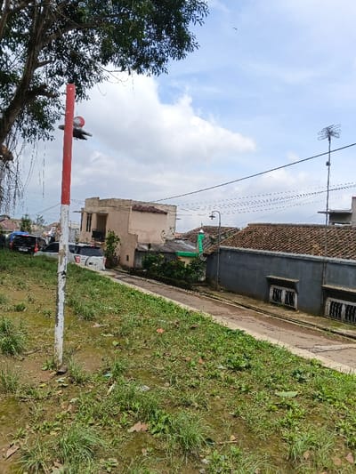 The image captures a daytime scene of a residential area with a sloping road, likely in Sarandi de Cuadra, Uruguay. In the foreground, a grassy and somewhat muddy embankment slopes downwards. A prominent red and white striped pole stands vertically on the left side, featuring a small, stylized bird-like ornament or weather vane near its top.
Behind the pole, on what appears to be a slightly higher section of the road, several vehicles are parked, including a dark and a white SUV. A narrow, paved road descends through the center of the image. Along the right side of the road, houses are visible. One house has a light-colored (possibly beige or pale orange) facade with multiple levels and a balcony. Further down the slope, another house features a dark grey wall with two barred windows and a tiled roof. A street lamp is positioned over the road in front of this house. Between the houses, a green structure with a dome or spire, resembling a small religious building or a unique architectural feature, is visible amidst some trees.
The upper left corner is dominated by the dark green foliage of a large tree, from which vines or aerial roots hang. The sky above is partially cloudy, indicating a bright day. In the background, power lines crisscross the scene, and a television antenna is mounted on a pole above one of the distant roofs. No specific activity or event is taking place, and no discernible text is visible on signs or vehicles.