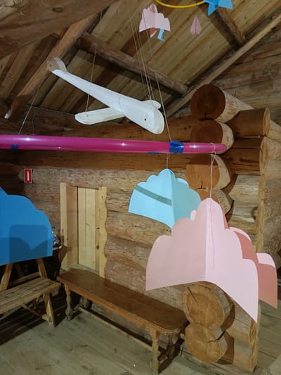 The image depicts the interior of a rustic wooden structure, likely a log cabin or a similar building, with a prominent sloped wooden ceiling and exposed log beams. Suspended from the ceiling are several decorative elements: a large white airplane model, a horizontal pink tube with blue tape wrapped around it, and several colorful paper cutouts resembling clouds and paper airplanes. These decorations suggest a playful or thematic setting, perhaps a children's area or an exhibition.
Below the hanging decorations, the walls are constructed from large, stacked logs. A small red sign with a white icon resembling a fire extinguisher is mounted on the log wall. In the foreground, there are two wooden benches: one shorter, simpler bench on the left, and a longer, more ornate bench on the right. The floor is made of wooden planks. Blue and pink cloud-shaped cutouts are also visible, hung on either side of the central space. The overall ambiance is warm and rustic, with the natural textures of the wood being a dominant feature. The scene is located in the city of Molochne, Russia.