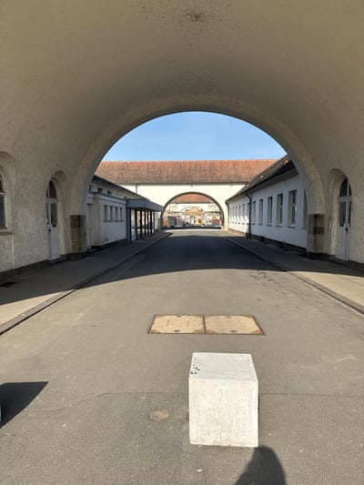 The image depicts a straight, paved passage viewed from within a large, light-colored arched entryway within the Sprudelhof complex in Bad Nauheim, Germany. The foreground features a light grey concrete block, roughly cubic, positioned slightly right of center on the asphalt path, casting a shadow to its right. A second partial shadow from another block is visible on the far left. A rectangular, two-part metal utility access cover is centered in the path just beyond the concrete block.
The passage extends forward, bordered by two rows of single-story, light-colored buildings with terracotta-tiled roofs. The building on the left exhibits several arched doorways, rectangular windows, and a section with dark-framed glass storefronts topped by a dark canopy. The building on the right has multiple rectangular windows and at least one arched doorway. Bright sunlight illuminates the scene, casting strong shadows from the overarching arch and the buildings onto the path.
In the mid-ground, a horizontal terracotta roof spans across the pathway, supported by another, smaller arched passageway through which the path continues. The background reveals a glimpse of further structures and indistinct architectural elements beyond this second arch. The sky above is clear and bright blue. No people are present.