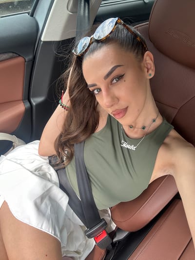 A young woman with long, dark hair is lying on her side in the passenger seat of a car. She is wearing a green tank top, white shorts, and a seatbelt. She has on sunglasses perched on her head, and a necklace with clover-shaped charms and her name, "Mariska," in script. On her left ear, she has a small, green clover stud earring. She is also wearing a bracelet on her left wrist with red, green, and black beads. The car has brown leather seats with a textured pattern. The interior of the car is visible, including part of the dashboard and seatbelt buckle. The image appears to have been taken during the daytime, with natural light illuminating the scene. The woman is looking towards the camera with a relaxed expression. The background shows the interior of a car.