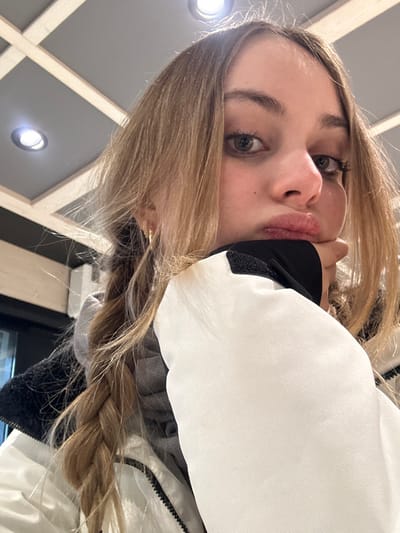 A close-up, rotated selfie shows a young woman looking directly at the camera. She has long, light brown hair, styled in a thick braid visible over her shoulder. Her light-colored eyes, possibly green or hazel, are prominent, and her lips are slightly pursed in a pouting expression. She is wearing a white winter jacket with black accents, and a black undergarment or glove is visible near her chin where her hand is positioned. The background, softly out of focus, reveals an indoor setting with a modern ceiling featuring a grid of light-colored wooden beams and several circular, bright recessed lights. The artificial lighting and the subject's winter attire suggest an indoor location, possibly a lodge or cafe, consistent with the given context of Bormio, Italy. No text is visible in the image. The overall impression is a casual, playful moment.