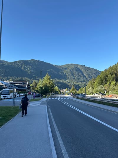 A person walks on a paved sidewalk next to a road in Kranjska Gora, Slovenia. The person is wearing a black shirt, black pants, and a backpack, and is carrying a gray bag. The sidewalk is bordered by a grassy area and a road with white lines. In the background, there are buildings with solar panels on the roofs, trees, and a large, forested mountain under a clear blue sky. Several buses and cars are parked along the roadside. The scene appears to be a sunny day.
