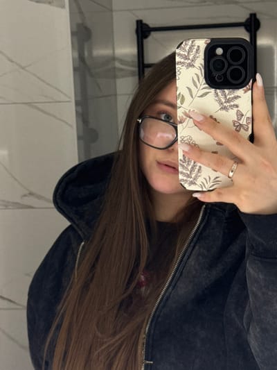 The image depicts a mirror selfie taken indoors in the city of Prikubanskii, Russia, likely within a bathroom or dressing area. A female individual is reflected from the chest up. She has long, straight, medium-brown hair and is wearing dark-framed glasses. Her left eye, with a blue iris, is visible. She is dressed in a dark, possibly charcoal grey or black, zip-up hooded jacket. Her right hand is holding an iPhone, which is equipped with a light-colored case featuring a botanical pattern of brown leaves, green foliage, and small butterflies. Her fingers display light-colored, manicured nails, and a thin silver-colored ring with an embedded design is present on her right ring finger. The phone partially obscures the right side of her face. The background consists of light-colored, possibly white or light grey, large-format tiled walls with subtle dark veining. A dark-colored, tubular heated towel rack is partially visible in the upper right quadrant of the reflection.