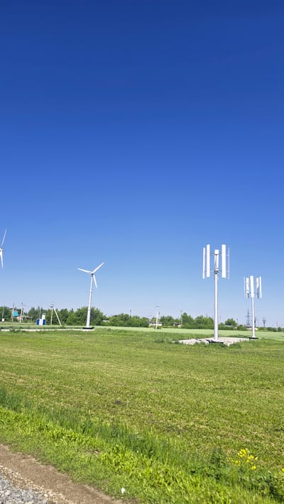 The image depicts a clear, bright day with a vast expanse of deep blue sky. In the foreground, a green, grassy field stretches across the frame, with a gravelly path visible on the lower left. In the mid-ground, several wind turbines are positioned within the field. To the left, a traditional three-bladed horizontal axis wind turbine stands tall. Further to the right, two smaller vertical axis wind turbines with multiple blades are prominent. A line of green trees forms a backdrop across the mid-ground, behind which a few buildings and power lines are discernible, suggesting a rural or semi-rural setting on the outskirts of a settlement. The overall scene conveys an impression of open space and renewable energy infrastructure. The location is confirmed to be the city of Актюбинский, Russia.