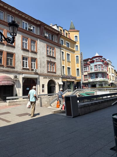 The image depicts a street scene in Plovdiv, Bulgaria, specifically on "Dom Bashtina Levski" street. In the foreground, a grey tiled walkway is partially covered by a shadow cast from the right. Two men are walking away from the camera. The man on the left, wearing a light blue polo shirt and tan pants, carries two white plastic bags. The man on the right, wearing a striped t-shirt and khaki pants, carries an orange bag. They are walking on a grey pathway towards a concrete ramp leading down.
Behind them, several buildings line the street. The building immediately behind the men is an older, grey structure with orange trim around its windows and architectural details. It features several windows, some with air conditioning units installed. An awning with the word "MIKA" is visible on the left side of this building. To its right, an archway leads into a building, and the words "DOM BASHTINA LEVSKI" are written above it.
Further down the street, a yellow building with a pointed green turret stands out. A vertical sign for "AURORA HILLS" is attached to its side, and below that, a sign for "AURORA HOTEL". Adjacent to the yellow building is a colorful, multi-story building painted in red, blue, and white, with balconies and decorative elements. Additional buildings of varying architectural styles and colors extend further down the street. The sky is clear and blue. The overall scene suggests a warm, sunny day in an urban environment.