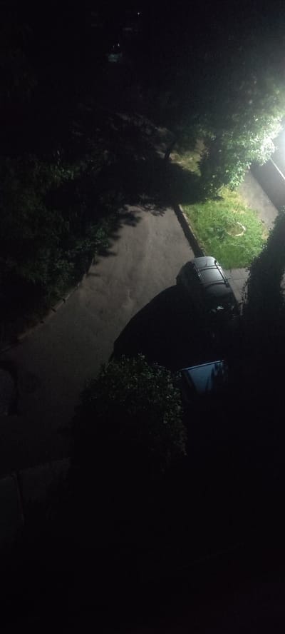 An aerial view captures a dimly lit outdoor scene at night, possibly in Svitlovodsk, Ukraine. A dark-colored car is parked on a paved surface. Adjacent to the car, a patch of green grass is visible, illuminated by a nearby light source. Dense foliage borders the paved area. In the background, the faint glow of lights suggests the presence of structures or streetlights. The overall impression is one of a quiet, urban or suburban environment at rest.