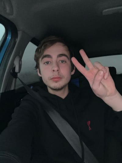 A young man poses inside a vehicle, wearing a black top with a small red logo. He is strapped in with a seatbelt. The man holds up a peace sign. He has light brown hair, a trimmed beard, and fair skin. The interior of the vehicle is visible, including a door and window. The photo appears to be taken in dim lighting.
