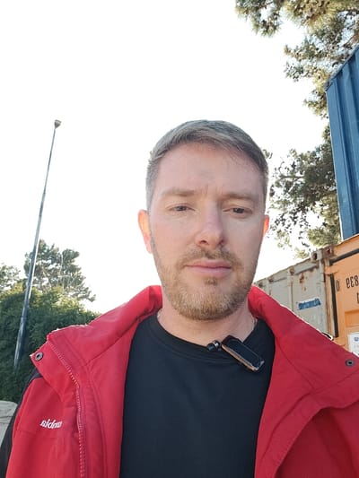 A fair-skinned male subject with light brown hair and a trimmed beard is depicted outdoors in Nesher, Israel. He faces the camera directly with a neutral expression, wearing a red zip-up jacket over a black crew-neck shirt. A black and brown electronic device, resembling a lapel microphone or Bluetooth earpiece, is clipped to his shirt. In the background to the left, a tall, grey street light pole extends vertically. To the right, a partial blue shipping container is stacked upon a full-height orange/tan shipping container, which displays faded white alphanumeric markings. Trees are visible in the background on both sides of the frame against a bright, overexposed sky.