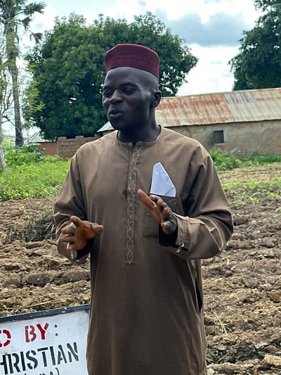 In the city of Afogo, Nigeria, a man stands in a field. He is wearing a brown robe and a red cap. His mouth is open as if he is speaking, and his hands are raised. There is a white cloth in his breast pocket. In the background, there is a building with a corrugated metal roof and trees. A sign in the foreground reads "BY: CHRISTIAN". The ground is covered in dirt and some sparse vegetation. The sky is cloudy.
