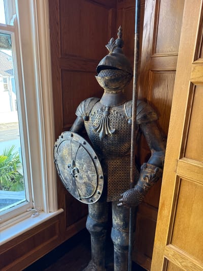 The image shows a full-length suit of armor positioned indoors, next to a window and a wooden door. The armor is made of dark metal with gold accents and features a shield and a spear. The window offers a view of a street with buildings and palm trees. The interior is decorated with wood paneling. The scene is static and appears to be a still life.