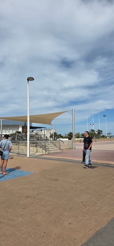 The image shows an outdoor scene in El Port De Sagunt, Spain. The sky is blue with scattered clouds. There are flags and palm trees in the background. There is a pedestrian area with light posts and shade sails. Two individuals are visible: a man in a black shirt and jeans and a woman in blue denim. They are walking on a paved walkway.
