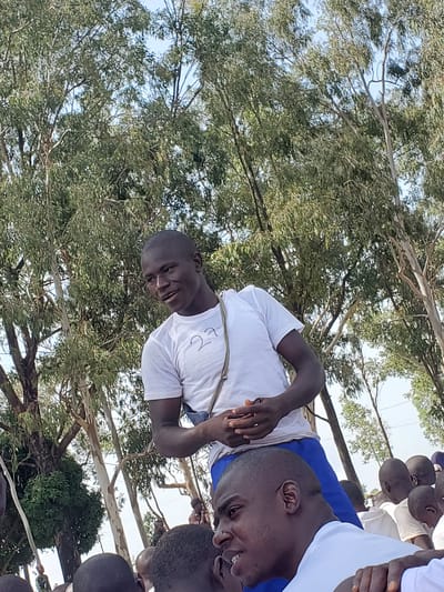 In Barakin Rafin Gora, Nigeria, a young man in a white t-shirt and blue shorts stands among a group of people outdoors. The background is dominated by tall trees with green foliage. The man is looking to his right and appears to be speaking. His hands are clasped together in front of him. The image captures a candid moment, with the man being the primary subject, and the surrounding environment and other individuals forming the context of the scene.