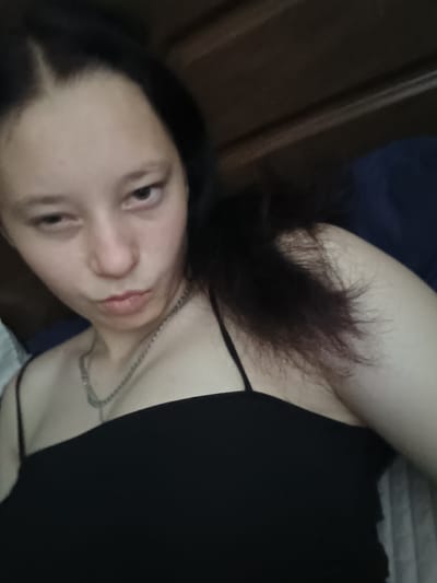 A young person with dark hair is looking at the camera with a pout. They are wearing a black tank top and a silver chain necklace. The background appears to be a bedroom, with dark wood furniture and a blue pillow. The image is a close-up selfie, and the lighting is dim. The image does not provide any information about the location in the city of Kalmykivka, Ukraine.