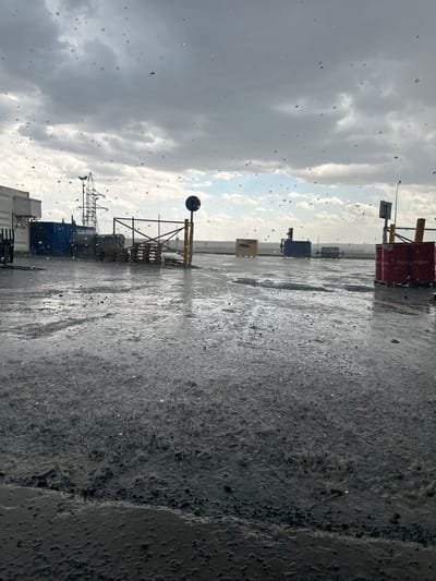 This is a rotated, eye-level, medium shot of a desolate industrial landscape under heavy rain. The foreground is dominated by a wet, gritty asphalt surface, reflecting the overcast sky. Scattered across the scene are various industrial objects, including large blue containers, a metal crane, and stacked crates, some of which are bright red. A yellow and black gantry-like structure with a white and black sign stands prominently in the middle ground, with a round, black sensor or light attached to a pole above it. Another similar pole with a light is visible further in the background. The sky is filled with heavy, dark gray clouds, interspersed with patches of lighter, brighter sky, suggesting a transition in weather. The presence of numerous raindrops on what appears to be a window or glass surface in the very foreground indicates that the photo was taken from inside a sheltered location looking out. The overall mood is somber and bleak, conveyed by the dark weather and the utilitarian nature of the setting. No discernible text is visible in the image.