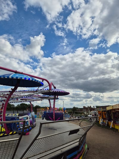 A Ferris wheel ride with spinning cars stands near other carnival rides at an outdoor fair or festival. The Ferris wheel features pink structural supports and blue passenger cars decorated with lights. The sky overhead is partially cloudy. The scene is likely in Babergh, United Kingdom.