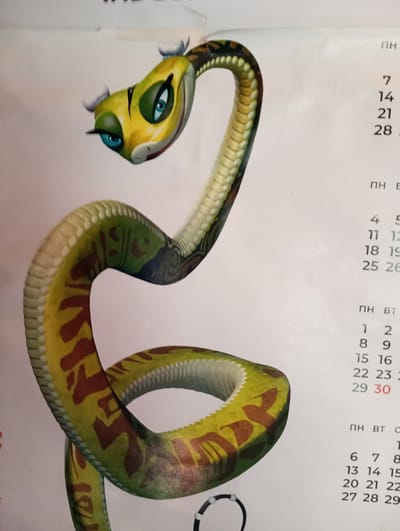 The image shows a cartoon snake character coiled on a white surface, which appears to be a calendar. The snake is predominantly green and yellow with brown markings. Its head is raised, and it has large blue eyes. The calendar has columns of numbers, suggesting it is displaying dates. The text "ПН" and "Вт" are visible, indicating days of the week. The provided crops do not offer any contextual information related to the city of Velykyi Sukhodil, Ukraine.