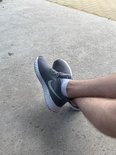 A pair of gray Nike running shoes with white soles and light gray socks are worn by a person whose legs are crossed. The shoes are positioned on a concrete surface that appears to be a sidewalk or pathway. The background consists of more concrete and a section of yellow-toned paving stones. The image provides a close-up view of the lower legs and feet, with the rest of the person's body not visible. The scene is outdoors, suggesting a casual setting.