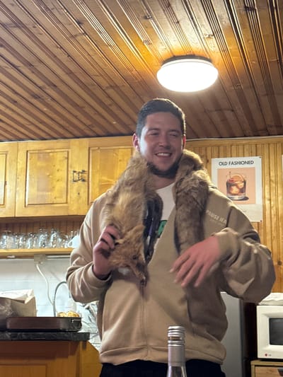 A man smiles while holding a fur stole in a room with wood paneling. A round light fixture is attached to the wall. A poster reading "Old Fashioned" is also on the wall, above wooden cabinets. A microwave is visible above a counter. There is no information in the image that places this scene in Lilkovo, Bulgaria.
