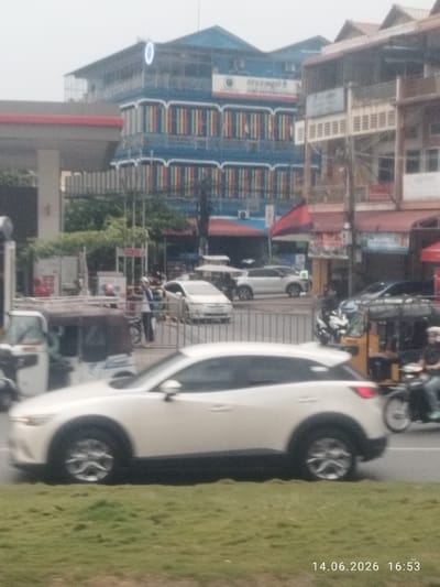 The image depicts a street scene in Khan Chbar Ampov, Cambodia, on June 14, 2026, at 16:53. In the foreground, a white car is captured in motion, with its front wheel and side clearly visible. Beyond the car, a street is lined with various buildings, some featuring colorful decorative elements and balconies. A gas station canopy is present to the left, indicating a commercial area. Several vehicles, including a tuk-tuk and motorcycles, are also part of the street traffic. People are present, some appearing to be walking or operating vehicles, suggesting everyday urban activity. The overall environment is a busy street with a mix of residential and commercial structures under an overcast sky.