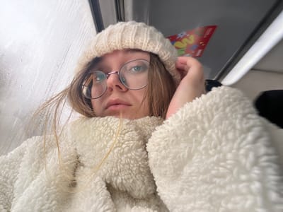 A low-angle, close-up image captures a light-skinned female individual indoors, positioned next to a window. Her light brown hair, reaching her shoulders, frames her face. She wears round, wire-rimmed glasses with light-colored eyes visible through the lenses, and a cream-colored ribbed knit beanie. Her expression is neutral, with her gaze directed upwards and slightly to her left. She is dressed in a thick, textured, off-white or cream-colored sherpa-style jacket or coat. Her left hand is raised near her head, partially touching her hat.

The window to her left is visible, covered in condensation or precipitation streaks, obscuring any exterior view. Above and behind her, to the upper right of the frame, a red object with yellow and green graphic elements, possibly a party hat or decorative item, is partially visible against a light grey overhead panel. The immediate environment suggests an interior space, likely within a vehicle such as a train or bus, or a room with a window, but a specific location within Борисово, Russia, cannot be identified from the visual evidence presented.