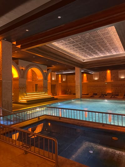 The image depicts a subterranean space in Ürgüp, Turkey. Visible architectural elements include stone walls with arched openings, a decorative ceiling with patterned tiles, and what appears to be a shallow pool or reflective surface with tiled edges. A metal railing and staircase are also visible. The space is illuminated with warm, artificial lighting, creating a yellow-orange ambiance. The scene is devoid of people.