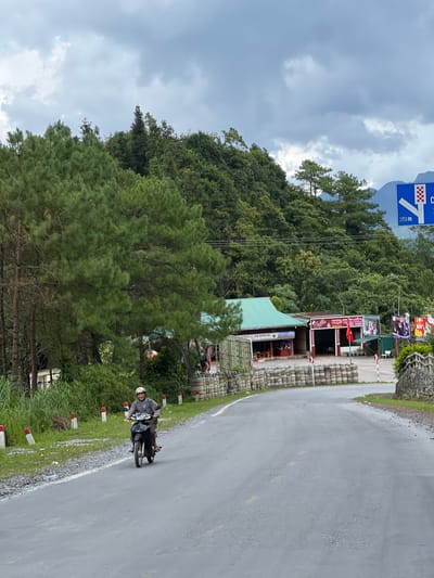 In Quản Bạ Commune, Vietnam, a man wearing a helmet rides a dark-colored motorcycle on a paved road. He is wearing a gray jacket and darker pants. The road curves to the right, and on the left side, there is a grassy verge with red and white bollards. Behind the grassy verge, tall evergreen trees and dense foliage cover a hillside.
On the right side of the road, there are several structures. One is a building with a green roof, and another, painted red and white, has signs with Vietnamese text and a red flag. A line of stacked tire barriers separates the road from these buildings. Further along the right side, there is a stone wall and more vegetation. A blue road sign with white symbols and the text "300m" is visible on a pole. The sky is overcast with gray clouds.