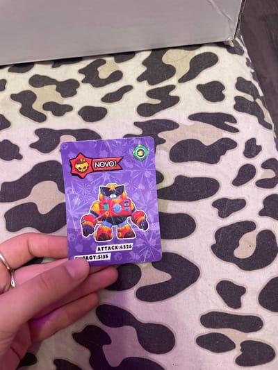 A person's hand, adorned with two silver rings on the index and middle fingers, holds up a purple trading card featuring a cartoon character with sunglasses and a red, orange, and yellow robotic body. The card is positioned over a patterned fabric with brown and beige splotches resembling a cow print. Text on the card reads "NOVO!" and "ATTACK: 4526". The background is out of focus, but a corner of a white cardboard box is visible in the upper portion of the image. The scene appears to be indoors, with artificial lighting casting a purple hue on the hand. The location is not identifiable as Vaganovo, Russia, or any specific city.