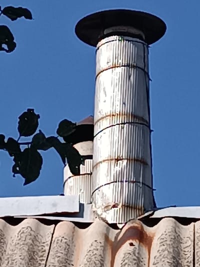 This media file shows a close-up view of a corrugated metal chimney with a dark cap, positioned on a similar corrugated roof. The chimney is cylindrical and appears to be constructed from vertically ribbed metal sheeting, with several black wires wrapped around it for support. There are visible signs of rust and wear on the chimney and roof, with reddish-brown streaks appearing to drip down the side of the chimney and onto the roof.
In the background, a clear blue sky is visible, and to the left of the chimney, dark green leaves from a branch extend into the frame. The lighting suggests it is daytime, likely sunny given the bright sky. The scene appears to be outdoors, possibly in a rural or suburban setting, given the materials of the roof and chimney. No people are visible in the image. There is no discernible text within the image. The overall impression is one of an aging but functional structure against a bright, clear sky.
