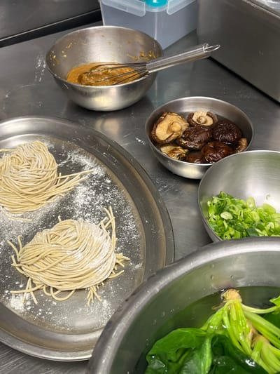 The scene depicts a detailed close-up of a food preparation area within a kitchen in Pozuelo de Alarcón, Spain. The primary surface is a stainless steel countertop, exhibiting minor smudges and reflections. In the immediate foreground, a portion of a large stainless steel bowl is visible, containing green leafy vegetables, consistent with spinach, partially submerged in clear water. To the left of center, a flat, round stainless steel tray holds two coiled bundles of raw, light-colored noodles, visibly dusted with white flour, with additional flour scattered on the tray surface. Adjacent to the noodles, in the midground, a medium stainless steel bowl contains several dark brown, rehydrated shiitake mushrooms submerged in a small amount of liquid. Next to the mushrooms, a smaller stainless steel bowl holds a quantity of finely chopped green onions. In the upper left background, another stainless steel bowl contains a viscous, light brown sauce, with a metal whisk resting diagonally across its rim. Further in the background, a light blue-lidded plastic container and other metallic kitchen equipment are partially visible. No individuals are present within the frame. The arrangement of ingredients suggests an active stage of meal preparation.