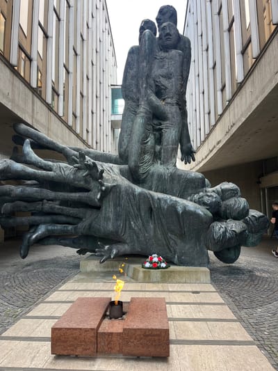 The image shows the Monument to the Slovak National Uprising in Banská Bystrica, Slovakia. It is a large, abstract bronze sculpture depicting a pile of bodies, with several figures reaching upwards. In front of the sculpture, there is an eternal flame burning in a rectangular stone structure, and a wreath of red and white flowers. The monument is situated in an outdoor plaza with cobblestone paving and a modern building with a glass facade in the background. A person in dark clothing is walking away from the monument on the right side of the image.
