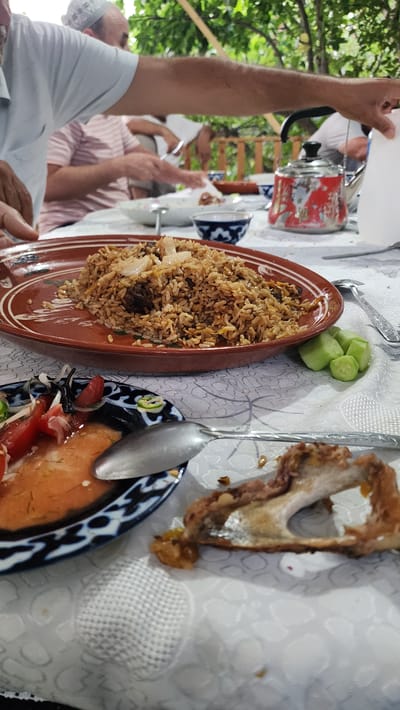This image depicts a meal scene outdoors, likely in Vodil, Uzbekistan, with a group of people gathered around a table laden with food. In the foreground, a large platter of pilaf, a traditional rice dish, is the central focus. To the left, a smaller plate contains a salad of tomatoes and onions, alongside a serving spoon. Further to the right, a piece of cooked meat bone rests on the table. In the background, several individuals are partially visible, engaged in eating. One person, wearing a white and patterned skullcap, is in the process of serving themselves. Another person, wearing a pink and white striped shirt, is also seated at the table. A red and white patterned teapot is visible in the middle ground. The setting appears to be a garden or outdoor dining area, with lush green foliage visible overhead, suggesting a warm, possibly summer day. The overall impression is of a communal meal shared in a relaxed, outdoor environment.