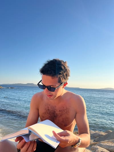 An outdoor scene captured in Agios Ioannis Diakoftis, Greece, depicts a shirtless male seated on a rocky shoreline reading a book under a clear blue sky. The individual, appearing to be a young adult, possesses dark, curly hair and moderate chest hair. He is wearing dark-lensed sunglasses with black frames and a watch with a light-colored band and a gold-toned, rectangular face on his left wrist. His left hand supports an open book with white pages and a light blue spine, while his right hand rests near the bottom right corner of the right page. He is looking down at the book. Behind him, calm, clear blue-green ocean water extends to a distant landmass on the horizon. In the mid-ground, near the left, dark rocks emerge from the water, with two faint human figures visible in the water nearby. The scene is brightly illuminated by natural sunlight, indicating a clear, sunny day.