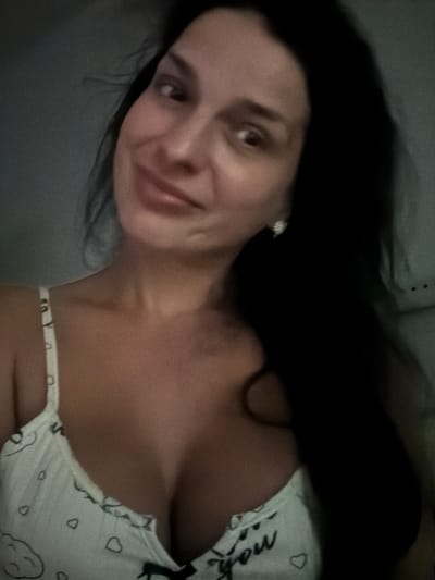 The image shows a medium shot of a woman with dark hair, wearing a white tank top with a heart pattern and the word "you" printed on it. She is looking towards the camera with a slight smile. The background is dark and indistinct. There are no identifiable landmarks or elements that would suggest a specific location within Lauderdale Lakes, Florida, or any other city.
