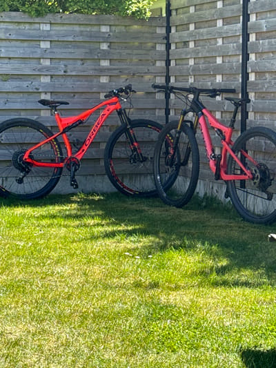 This image captures two mountain bikes prominently displayed outdoors against a wooden fence, with green grass to the right. The scene is bathed in bright daylight, indicated by strong shadows cast across the grass and fence, suggesting it is mid-morning or afternoon on a sunny day.
The primary subjects are two full-suspension mountain bikes, both identified by the "ORBEA" branding visible on their frames. The bike in the foreground is a vibrant orange, with black accents and gold-colored suspension components. Its tires, branded "MAXXIS," are black with aggressive tread patterns, and its pedal, chainring, and rear cassette are clearly visible. The second bike, positioned behind and slightly above the orange one, is a distinct pink or salmon color, also featuring black components and disc brakes. "WOSSOV" is faintly visible on its tire. Both bikes are propped up, likely on a stand or leaning against a fence, though the support structure is partially obscured by the bikes and the fence.
The background consists of a tall wooden fence, made of vertical planks and horizontal beams, which runs across the left side of the frame. To the right of the bikes, a lush green lawn or grassy area extends into the distance. No people are present, and no specific activity is taking place other than the bikes being stored or displayed. The overall impression is one of an outdoor setting, potentially a backyard, with the bikes ready for use. While the location context is given as Bredene, Belgium, no specific visual cues within the image confirm this geographical detail.