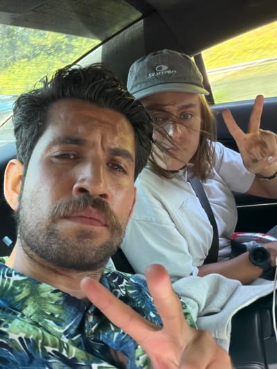The media file is a selfie taken inside a vehicle. Two people are visible: one in the foreground and another in the background. The person in the foreground, who appears to be male, has dark, wavy hair and a beard. He is wearing a patterned shirt with blue and green leaves. His right hand is raised, with two fingers extended, forming a peace sign.
The person in the background, appearing to be female, is wearing a grey baseball cap with the word "SKATEPRO" visible on it. She is wearing a white shirt and also has her right hand raised, forming a peace sign. Her hair is light brown and windswept. Both individuals seem to be looking at the camera.
The background of the image shows a blurred view of trees and greenery through a window, suggesting the vehicle is in motion, possibly on a road trip. The lighting indicates it is daytime. There is a seatbelt visible across the person in the background. The overall mood appears to be casual and cheerful, with the peace signs suggesting a fun or friendly interaction. No specific time of day or weather conditions can be definitively determined, though the bright lighting implies good weather. The location context provided is Clamart, France.