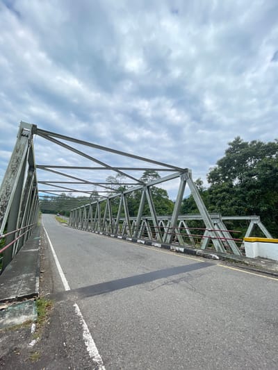 The image shows a view from the side of a bridge with a metal truss structure. The bridge has an asphalt road with white lines. On the right side of the road, there are some yellow and white concrete barriers. Lush green trees are visible beyond the bridge. The sky is cloudy. The scene appears to be in a rural or semi-rural area.