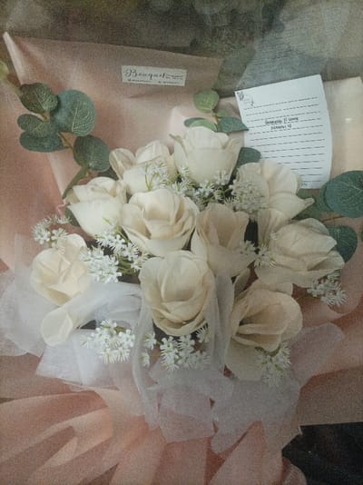 This image presents a detailed, static view of an artificial flower bouquet, located in Pasir Muncang, Indonesia. The bouquet consists primarily of multiple cream-colored artificial roses, interspersed with small white artificial filler flowers and green artificial foliage resembling eucalyptus leaves. It is wrapped in a two-layered packaging: an inner layer of gathered sheer white fabric and an outer layer of light peach-pink paper. Visible on the peach wrapping in the upper left is a small label with the text "Bouquet by Novia" and associated social media handles. A white, lined note card bearing handwritten text, including "Barakallah fi Umur" and "MAMPU! <3", is positioned on the upper right side of the bouquet. The scene is illuminated by soft, diffused light, indicating an indoor environment. No individuals are present, and no actions or interactions are depicted.
