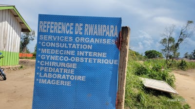 This outdoor image captures a blue metal sign detailing medical services offered at a "REFERENCE DE RWAMPARA" facility in Tchere, Democratic Republic of the Congo. The sign, made of diamond plate metal, is partially obscured on the right by a concrete post and shows signs of wear, with some reddish material near its attachment point.

The text on the sign, in white capital letters, lists:
- "REFERENCE DE RWAMPARA"
- "SERVICES ORGANISES" (Organized Services)
- "*CONSULTATION"
- "*MEDECINE INTERNE" (Internal Medicine)
- "*GYNECO-OBSTETRIQUE" (Gynecology-Obstetrics)
- "*CHIRURGIE" (Surgery)
- "*PEDIATRIE" (Pediatrics)
- "*LABORATOIRE" (Laboratory)
- "*IMAGERIE" (Imaging)

In the background to the left, a small, white wooden building with a green roof trim is visible, alongside what appears to be part of a motorcycle. The foreground features a dirt road and abundant green vegetation, indicating a rural or semi-rural setting. To the right, another white sign lies face-down in the grass. The sky is partly cloudy, suggesting daytime. Trees and more vegetation populate the distant background under a bright, diffused light. The bottom right corner of the image contains a watermark with "Joseph Kisuki".
