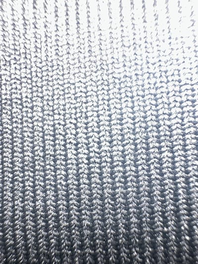The image displays a close-up view of a textured, knitted fabric, likely a textile material. The fabric exhibits a repeating pattern of interlocking stitches, creating a chevron-like or herringbone design. The color is predominantly silver or metallic gray, with variations in light and shadow suggesting a subtle sheen or reflective quality of the material. The texture is finely detailed, with individual threads or loops visible.

There is no discernible location within the city of Letterkenny, Ireland, depicted in this image. The image focuses solely on the fabric's texture and pattern, lacking any contextual elements that would identify a specific place. No people, subjects, objects, actions, or interactions are present in the scene. The environment is abstract, reduced to the surface of the fabric itself.