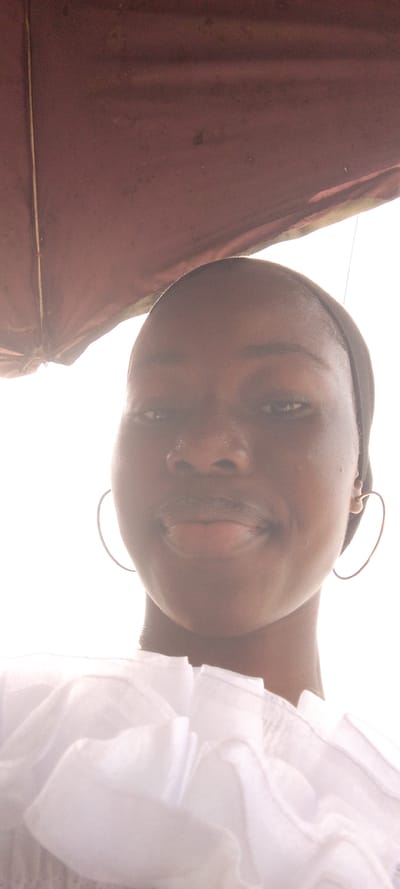 A low-angle, overexposed portrait captures a female subject, estimated to be in her late teens or early twenties, in Ohokobe Ndume, Nigeria. The subject, possessing a dark skin tone, dark eyes, and full lips, displays a slight, closed-mouth smile while looking directly into the camera. She wears a dark head covering and large, thin, metallic heart-shaped hoop earrings. The visible portion of her attire is a white garment featuring ruffled or pleated fabric around the upper chest. Directly above the subject, occupying the top quarter of the frame, is a reddish-brown, weathered fabric structure, consistent with a canopy or umbrella, exhibiting a vertical light-colored seam on its left side. The remainder of the background is overexposed to white due to bright ambient lighting.