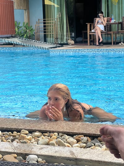 This image depicts an outdoor swimming pool area in Ban Tha Muang, Thailand, under daylight conditions. In the midground, a fair-skinned female with wet, light-colored hair is partially submerged in the blue-tiled pool, resting her upper torso on the pool's edge. She wears a dark-colored swimsuit, and her hands are clasped near her mouth while she gazes towards the upper right of the frame. Reddish marks are visible on her cheeks, and a white earring is present on her left ear.
In the background, on a tiled patio adjacent to the pool, a second female individual with dark hair is seated on a wooden chair at a wooden table. She is dressed in a dark top, a white skirt, and dark flip-flops, and is actively engaged with a smartphone held in both hands. A plastic water bottle is positioned on the table beside her. Behind the patio, a building with a large reflective glass door is visible, along with green foliage and a wooden privacy fence on the left. A bamboo rack stands upright near the building.
A black mesh hammock-style lounger is partially visible, extending from the pool edge towards the background on the left. The pool coping in the foreground consists of a light brown stone slab bordered by embedded pebbles. In the extreme foreground, on the bottom right, the bare toes and front portion of a human foot are visible, resting near the pool's edge.
