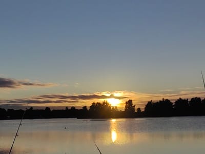 The image shows a tranquil sunset over a lake, likely in the Усяжскі сельскі Савет region of Belarus. Silhouetted against the colorful sky are trees lining the distant shore, with a few indistinct clouds present. The sun, a bright orb, is partially obscured by clouds and trees, casting a shimmering reflection on the water's surface. In the foreground, two fishing rods are visible, angled towards the water, suggesting a recreational fishing activity. The overall scene evokes a sense of calm and natural beauty.