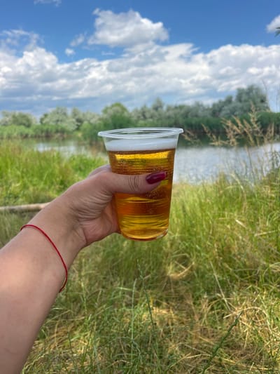 A person's hand with a red bracelet and burgundy nail polish holds a clear plastic cup filled with beer. The beer is golden yellow with a frothy white head. The background is a blurry natural scene with a body of water, green grass, and trees under a bright blue sky with white clouds. The image appears to be taken outdoors during the daytime. There is no visible text in the image. The overall impression is one of relaxation and enjoyment in a natural setting.