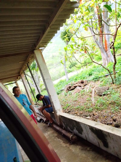 In Atamo Sur, Venezuela, two men are observed under a covered walkway. To the left, a man wearing a bright blue t-shirt and red shorts stands facing forward, looking away from the camera. To his right and further back, another man in a dark blue t-shirt and shorts sits on a concrete ledge, looking down at his phone. The walkway's corrugated roof is visible overhead, supported by concrete pillars. Beyond the walkway, a lush, green hillside slopes downwards, dotted with trees and vegetation. The ground beneath the walkway appears to be concrete, with a pipe running along the ledge. The overall scene depicts a casual, outdoor setting with a natural backdrop.