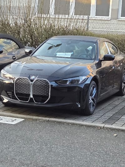 Location: Sprendlingen, Germany.
The image presents a stationary black modern BMW coupe, likely a 4 Series model, parked on a surface of grey rectangular pavers. The vehicle faces slightly left, displaying its prominent vertical kidney grille with a textured mesh design, integrated headlights, and multi-spoke alloy wheels. A white document is visible on the dashboard through the windshield. Partially visible to the left and rear of the BMW is a dark-colored convertible vehicle with its soft top retracted. The paved parking area transitions to an asphalt road in the foreground, separated by a grey curb. A white numerical marking, "_3", is partially visible on the asphalt. The background features a dense line of bare deciduous vegetation, behind which a light-colored building with multiple white-framed windows and a horizontal fence is situated. The scene is illuminated by natural daylight. No individuals are visible.