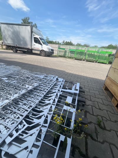 The image depicts an outdoor industrial or logistics area in Bogucin, Poland, on a sunny day with a blue sky and scattered clouds. In the foreground, a large stack of thin, laser-cut metal sheets with intricate geometric patterns is laid out on a wooden pallet. Some yellow wildflowers are growing amidst the metal sheets. To the left, a white box truck with a grey tarp-covered trailer is parked, bearing the logo "OSS MET" on its side. In the background, a row of bright green shopping carts or trolleys is lined up, some with visible numbering such as "089". Beyond the carts, a brick wall and trees are visible, further suggesting an outdoor facility. The ground is paved with interlocking concrete pavers. No people are present in the scene.