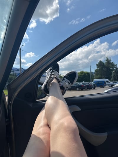 A person's legs are extended out of a car window, with their feet wearing black and white Vans sneakers. The car is parked in a lot with other vehicles, including a blue van, and trees and buildings are visible in the background under a blue sky with white clouds. The image was taken in the city of Iwye, Belarus.