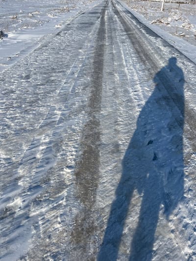 An upward-angled view captures an icy, snow-covered dirt or gravel road in Qoraobdol QFY, Uzbekistan, under clear, sunny conditions. The road, running centrally and receding into the distance, features multiple parallel ruts and tracks formed by vehicles in the frozen surface. Patches of darker, clearer ice are interspersed with textured snow and ice. To the left and right of the road, the surrounding terrain is blanketed in snow, with sparse, low, dry vegetation visible through the white covering. A prominent, elongated human-like shadow, cast towards the upper left from the lower right foreground, occupies the right half of the image, with discernible foot-shaped depressions within the shadow on the snow. The sun appears to be low in the sky, illuminating the scene from behind the observer, who is presumably the source of the shadow.